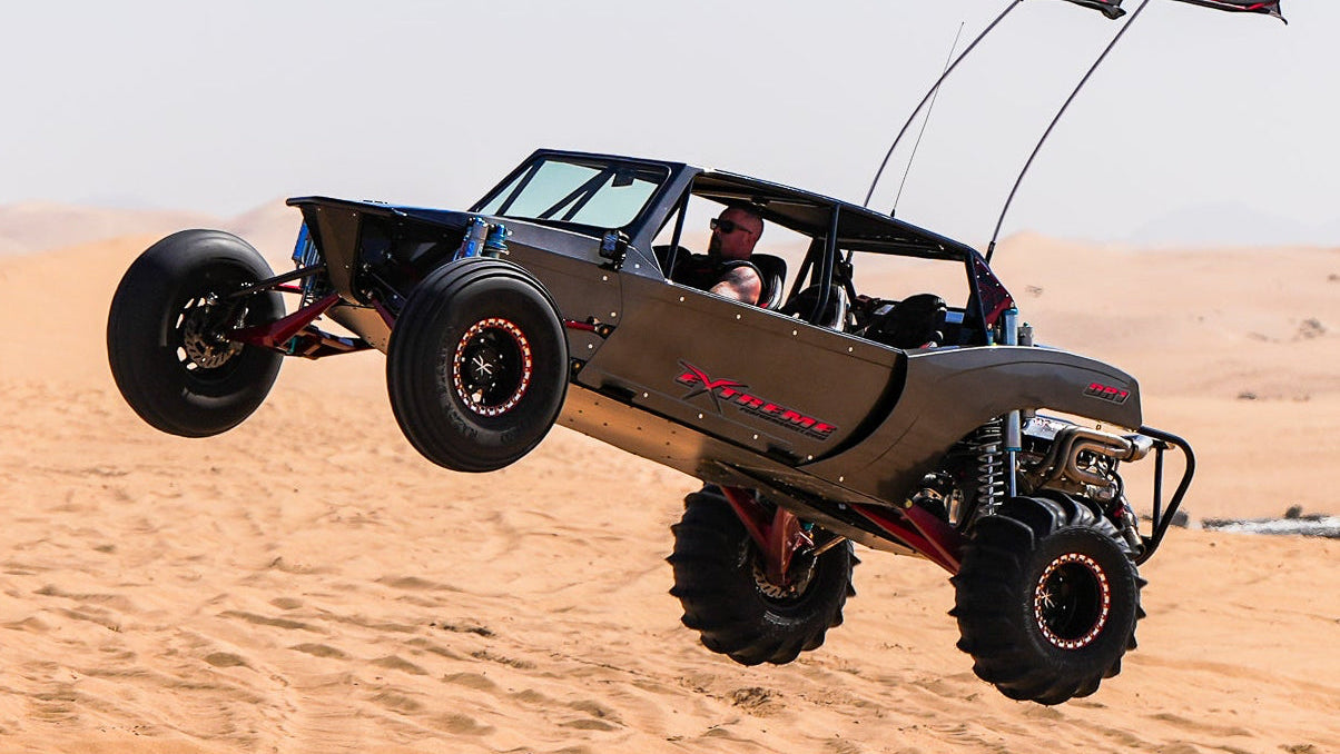 A desert sand buggy jumps high into the air over sandy dunes, with two small flags attached to the rear antennas. The vehicle is shown in mid-air against a clear blue sky and desert landscape.